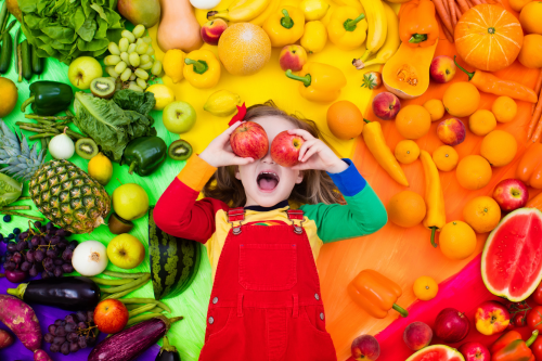 Little girl smiling and holding apples over her eyes laying on a variety of colorful, healthy fruits and vegetables
